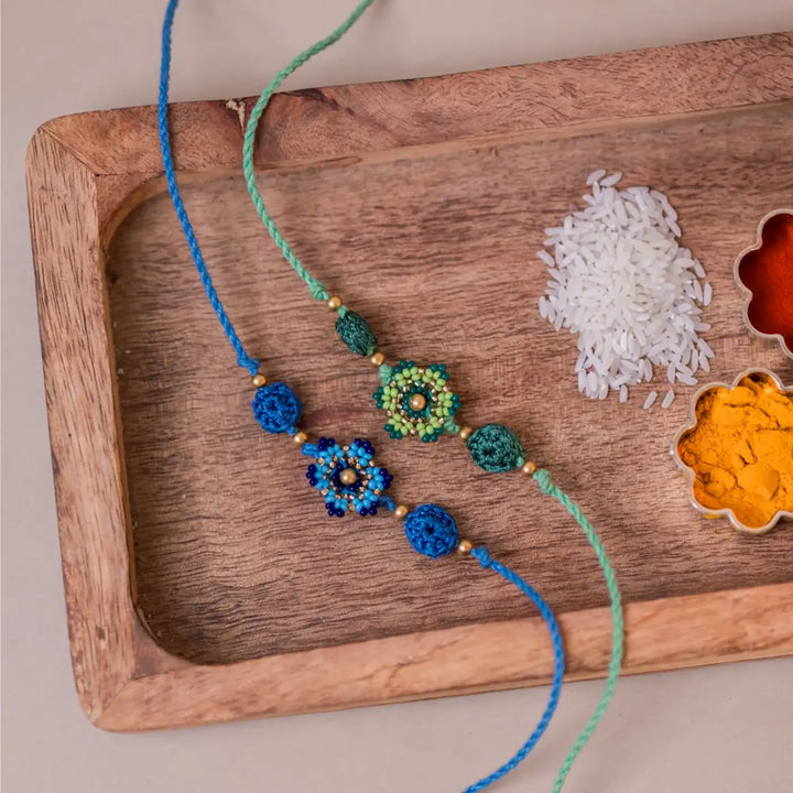 Handmade Crochet Blue And Green Flower Rakhi With Roli Chawal Set Of 2 - Top View in Wooden Tray