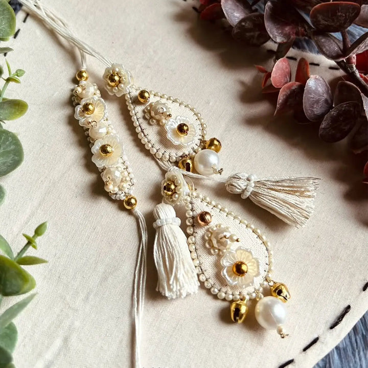 Hand Embroidered Beaded Ivory Bhaiya Bhabhi Rakhi With Roli Chawal Set Of 2 - Close-up of Earrings on Fabric
