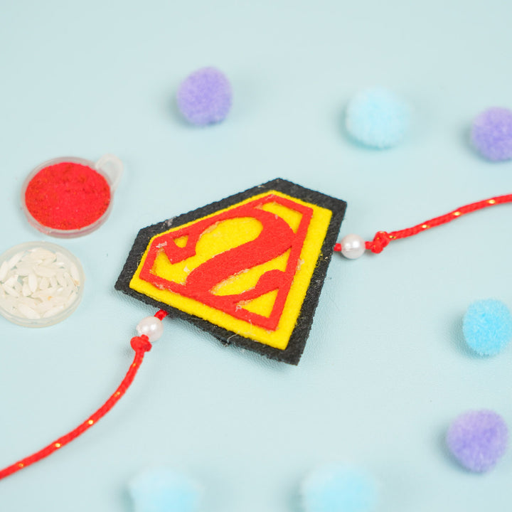 Handmade Beaded Superman Theme Pom Pom Felt Rakhi With Roli Chawal For Kids - Flat lay with Rakhi and festive items