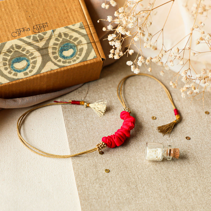 Handmade Reusable Gold Beads And Gathered Upcycled Fabric Rakhi With Roli Chawal For Brother - Flat Lay with Box and Bottle