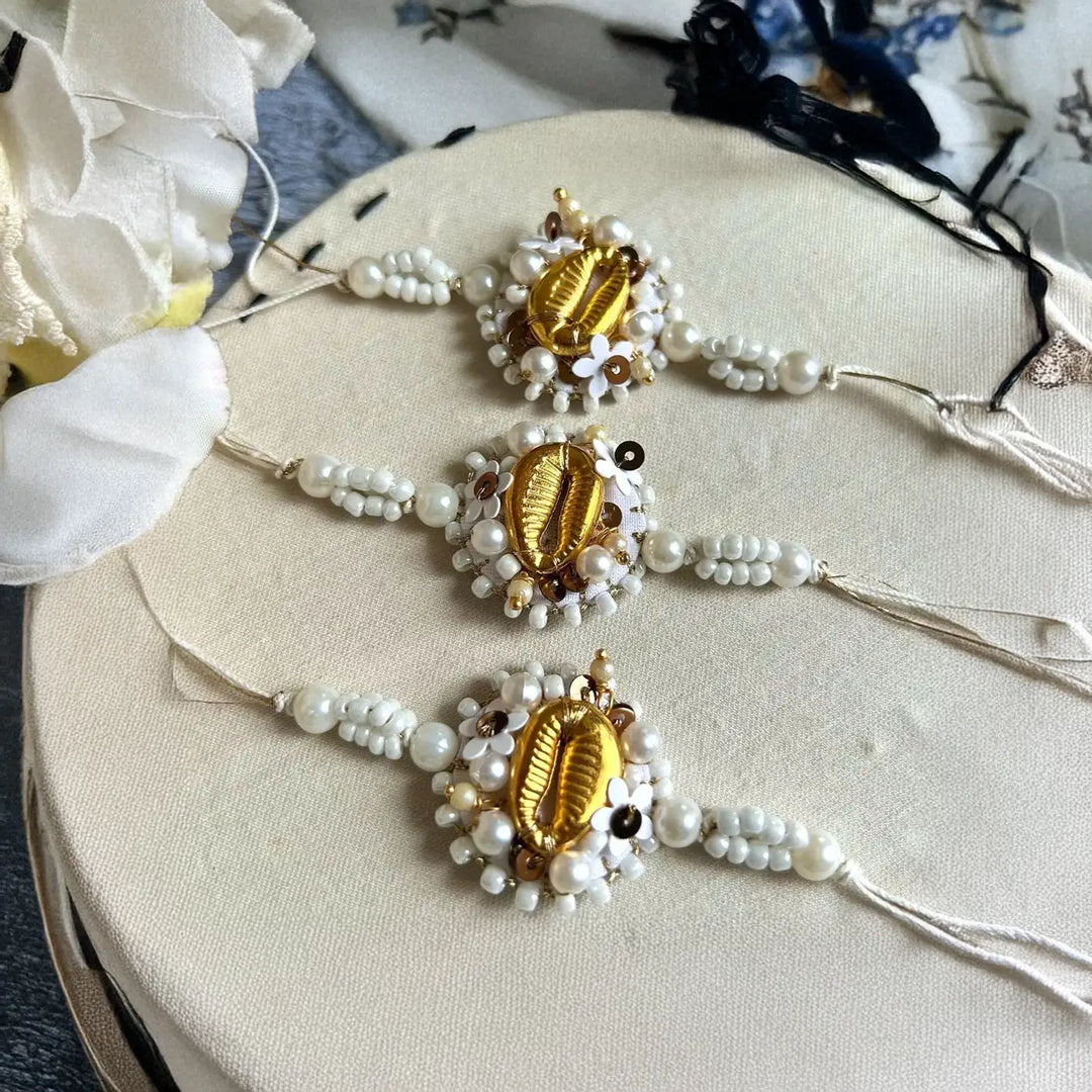 Hand Embroidered Beaded White And Golden Shell Rakhi With Roli Chawal Set Of 3 - Three Bracelets Laid on Round Surface