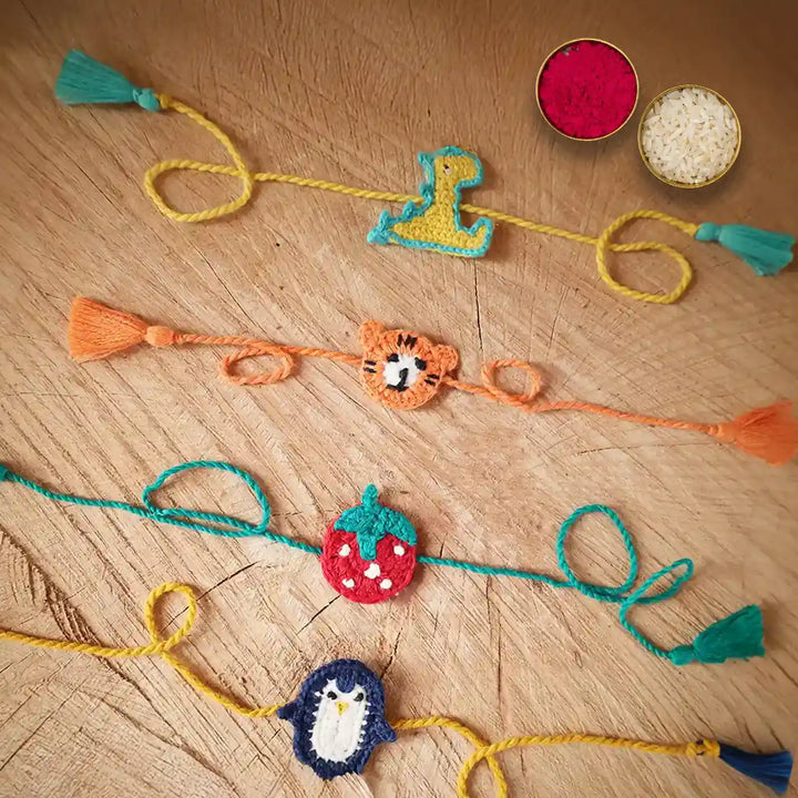 Handmade Crochet Dino Tiger Strawberry And Penguin Kids Rakhi With Roli Chawal Set Of 4 - Flat Lay with Ritual Accessories