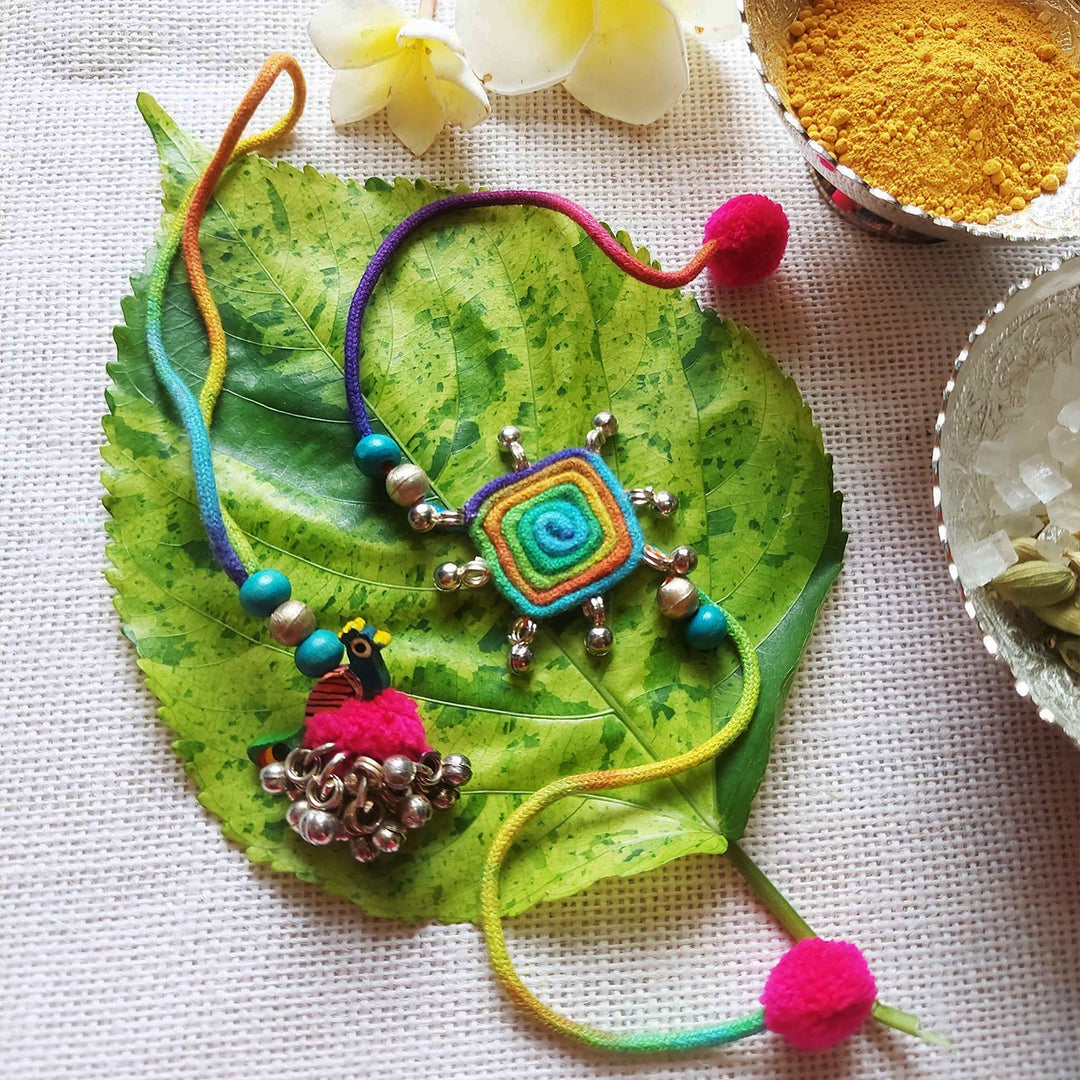 Handmade Dori Work Peacock And Ghungroo Bhaiya Bhabhi Rakhi With Roli Chawal Set Of 2 - Ornaments displayed on green leaf