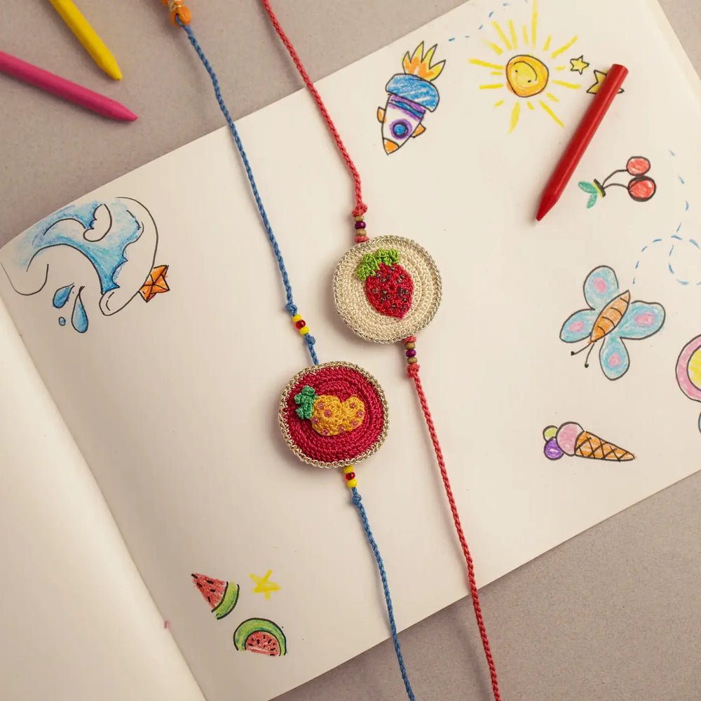 Handmade Crochet Pineapple And Strawberry Theme Kids Rakhi With Roli Chawal Set Of 2 - Flat Lay on Open Notebook