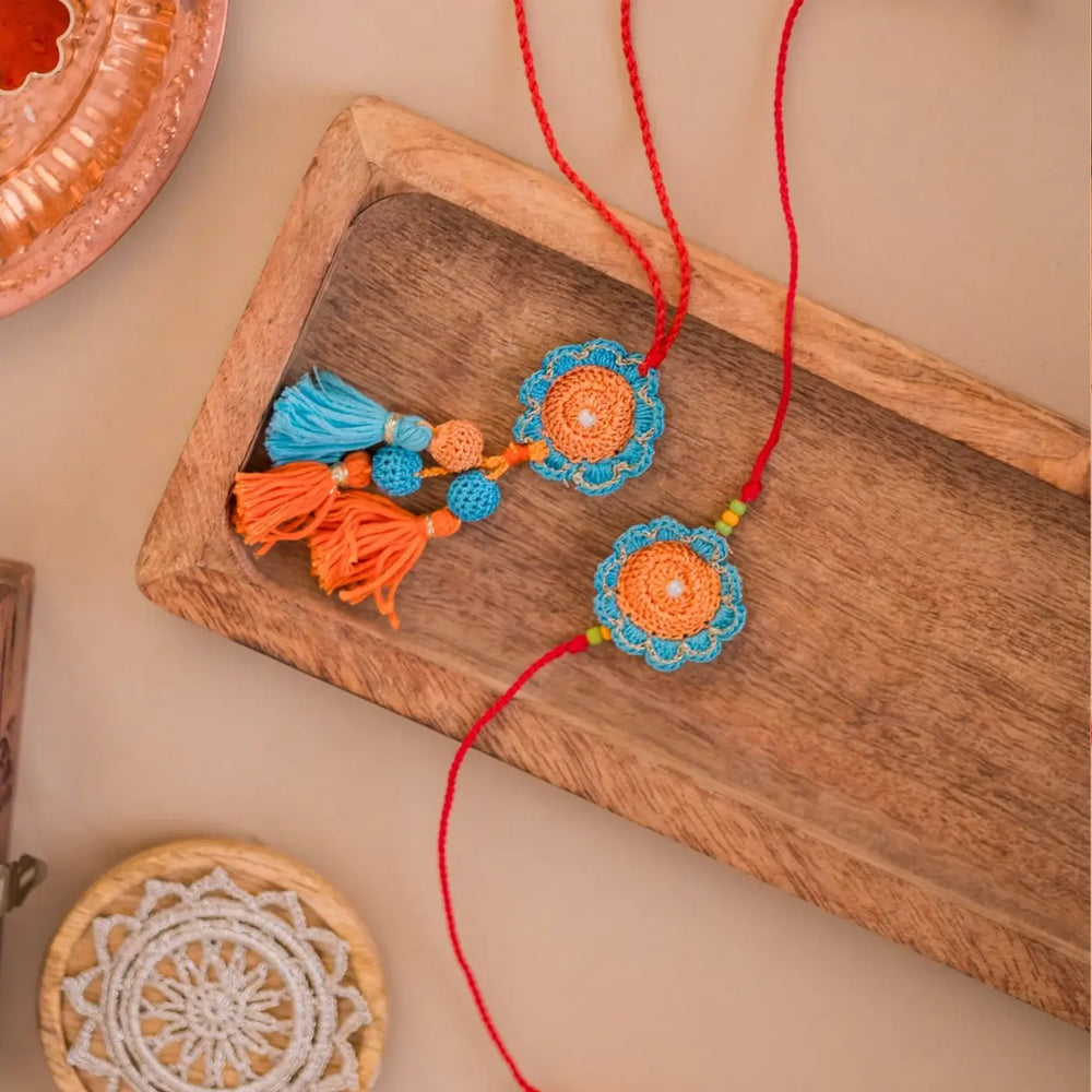 Handmade Crochet Blue And Orange Camelia Bhaiya Bhabhi Rakhi With Roli Chawal Set Of 2 - Overhead View with Background Accents