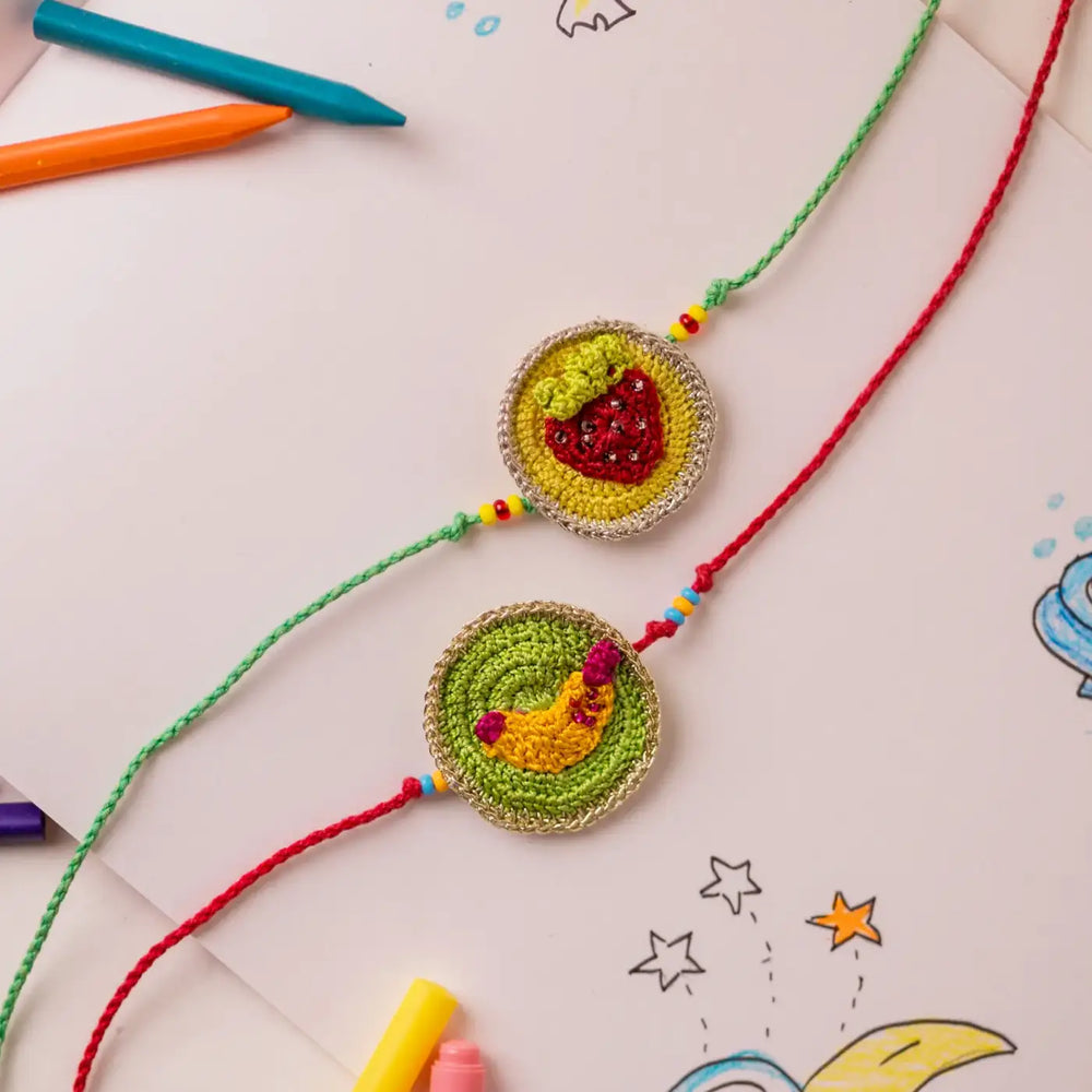 Handmade Crochet Banana And Strawberry Theme Kids Rakhi With Roli Chawal Set Of 2 - Flat Lay with Drawing Background