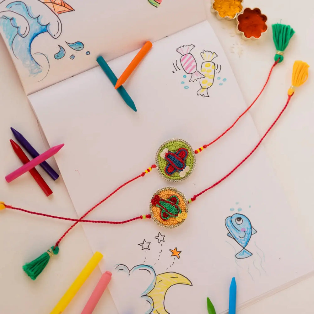 Handmade Crochet Airplane Theme Kids Rakhi With Roli Chawal Set Of 2 - Flat Lay with Drawing Accessories