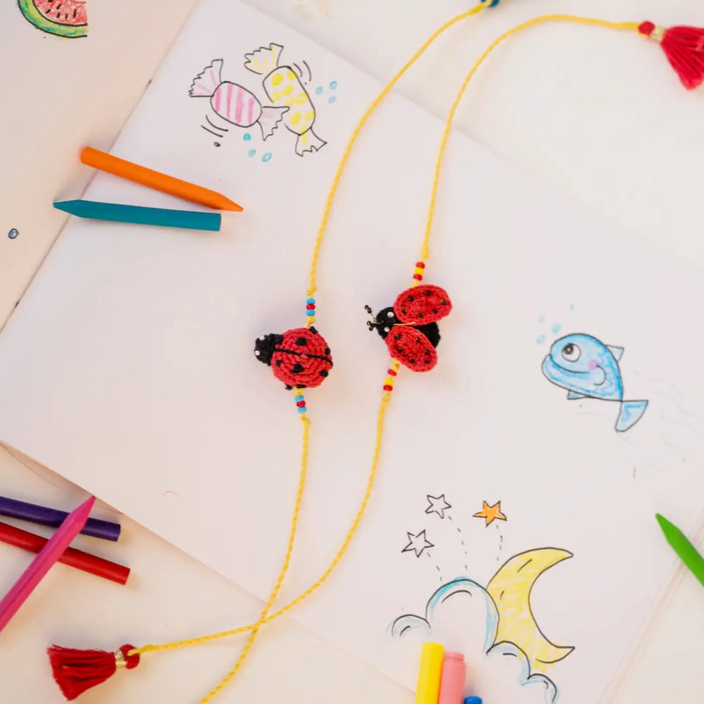 Handmade Crochet Mood Beetles Theme Kids Rakhi With Roli Chawal Set Of 2 - Laid Flat with Background Drawing