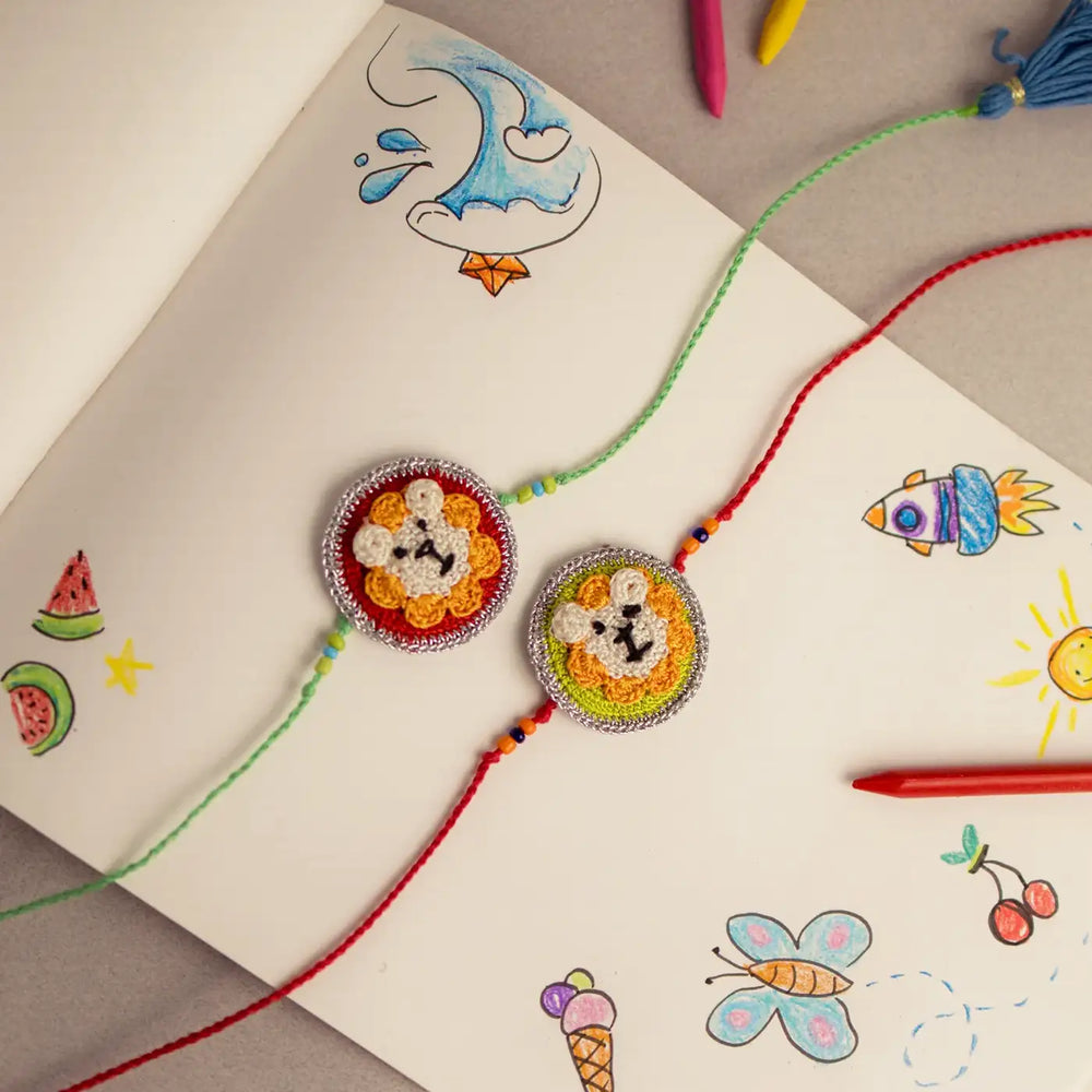 Handmade Crochet Lion Theme Kids Rakhi With Roli Chawal Set Of 2 - Overhead View with Sketchbook Background