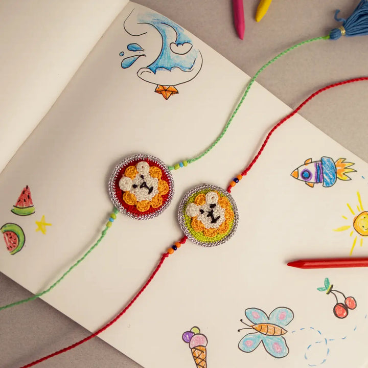 Handmade Crochet Lion Theme Kids Rakhi With Roli Chawal Set Of 2 - Overhead View with Sketchbook Background