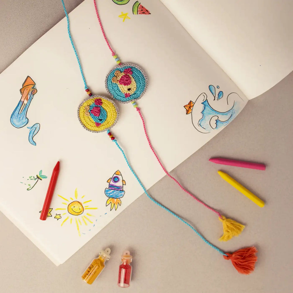 Handmade Crochet Bird Theme Kids Rakhi With Roli Chawal Set Of 2 - Laid on Sketchbook with Crayons