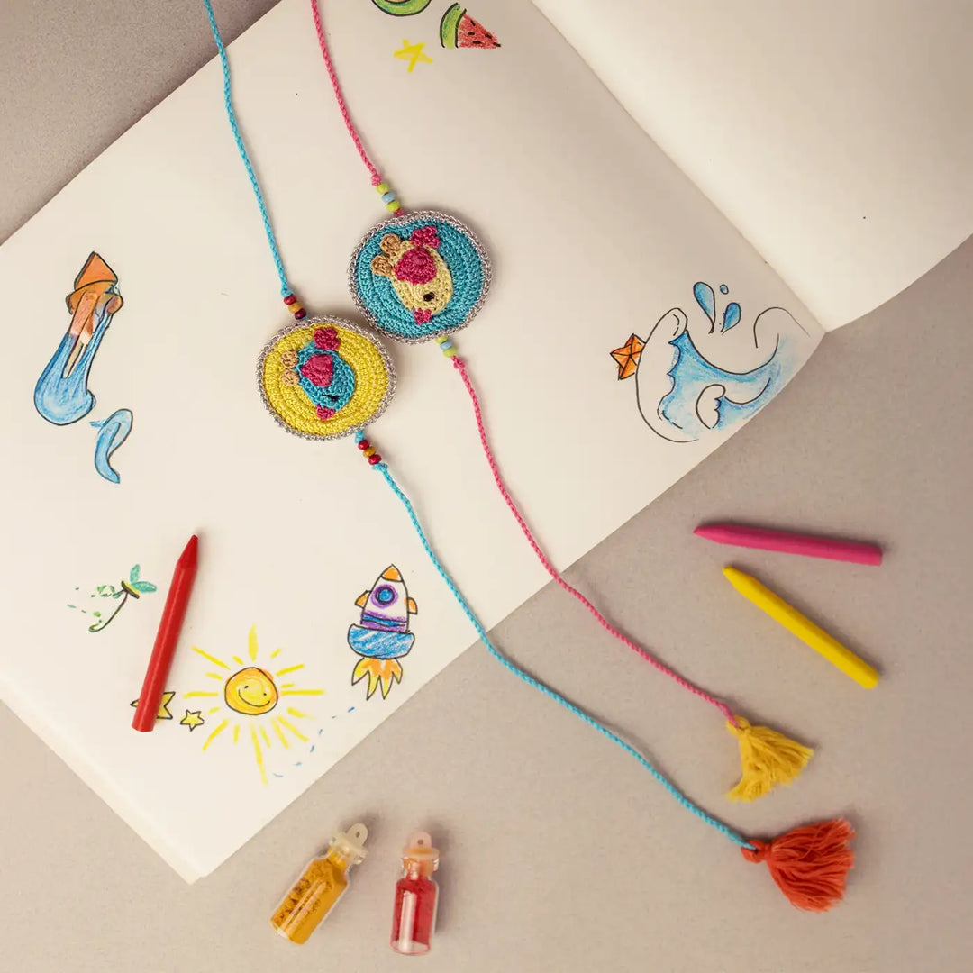 Handmade Crochet Bird Theme Kids Rakhi With Roli Chawal Set Of 2 - Laid on Sketchbook with Crayons