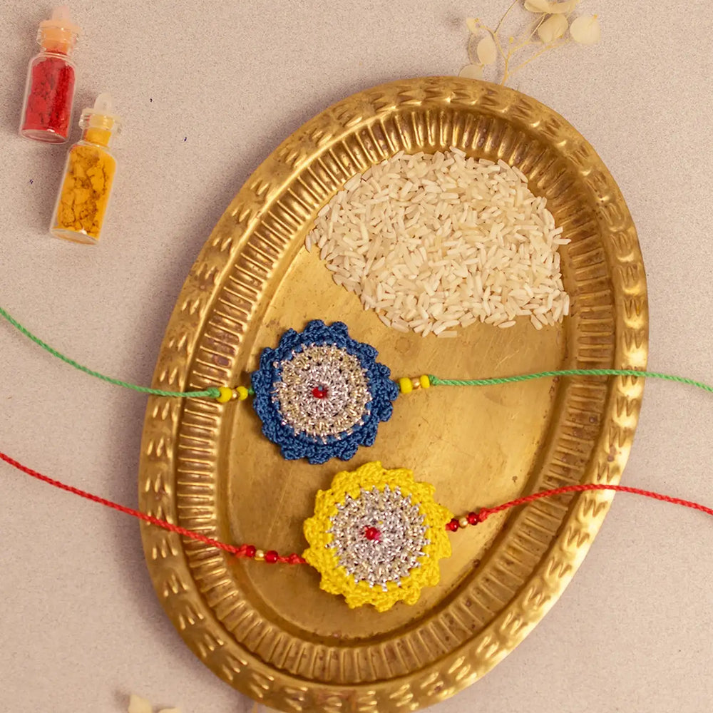 Handmade Crochet Blue And Yellow Seher Rakhi With Roli Chawal Set Of 2 - Top-down view with rice and colors