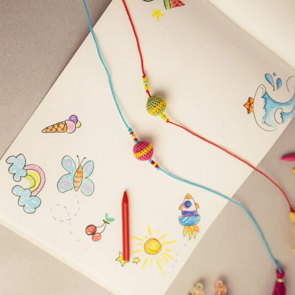 Handmade Crochet Multicolor Suraj Rakhi With Roli Chawal Set Of 2 - Laid on Sketchbook with Drawings