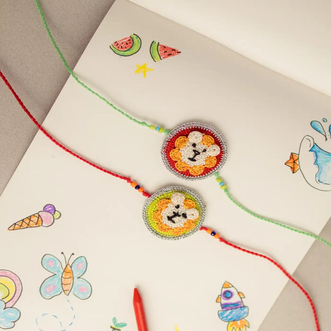 Handmade Crochet Lion Theme Kids Rakhi With Roli Chawal Set Of 2 - Top View with Sketchbook and Crayon