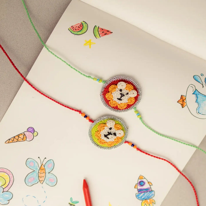 Handmade Crochet Lion Theme Kids Rakhi With Roli Chawal Set Of 2 - Top View with Sketchbook and Crayon