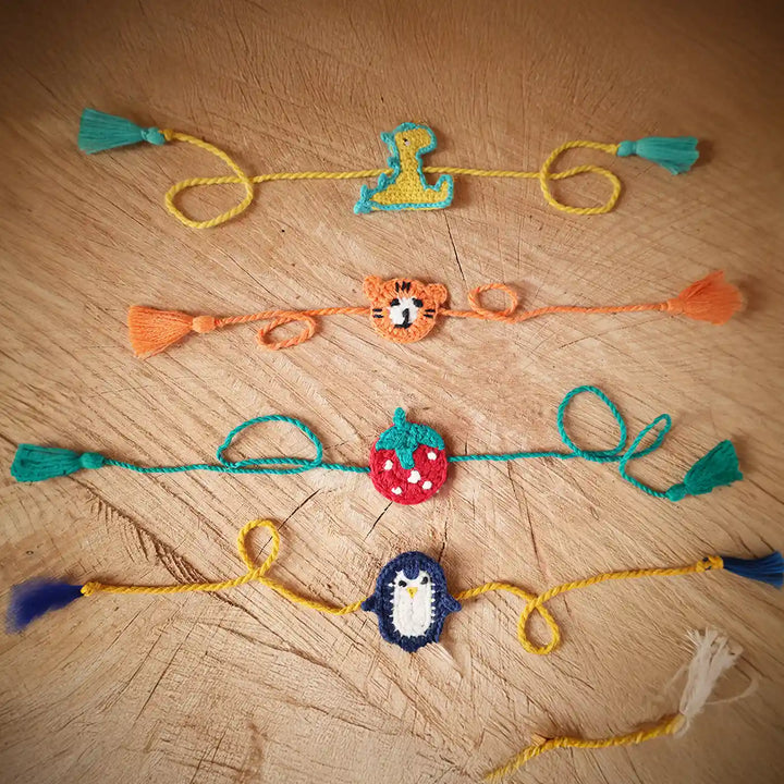 Handmade Crochet Dino Tiger Strawberry And Penguin Kids Rakhi With Roli Chawal Set Of 4 - Flat Lay of Bracelets with Tassels
