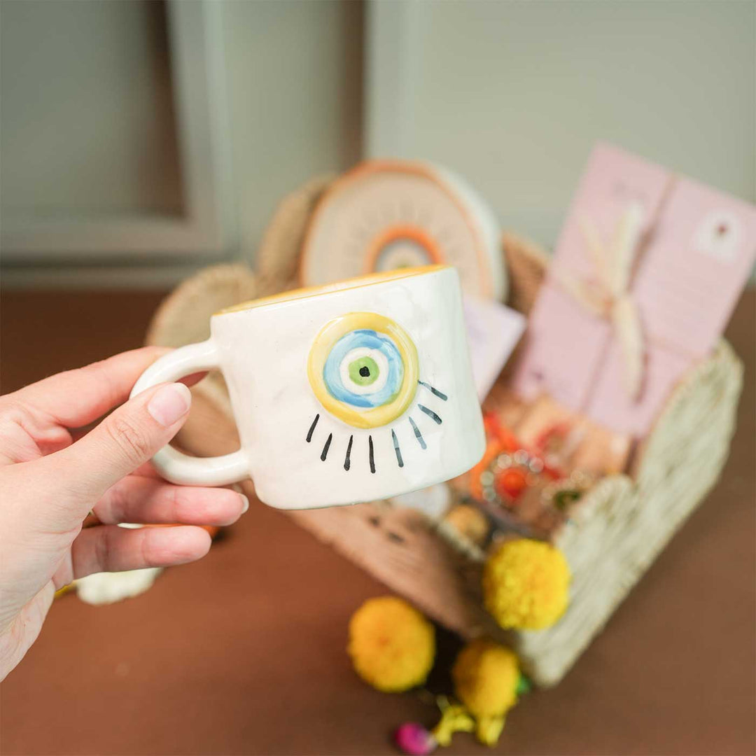 Personalized Handmade Evil Eye Design Guardians Blessing Rakhi Hamper Includes 1 Basket 1 Cup And Saucer 1 Rakhi 1 Roli Chawal And 1 Message Card - Hand holding ceramic cup