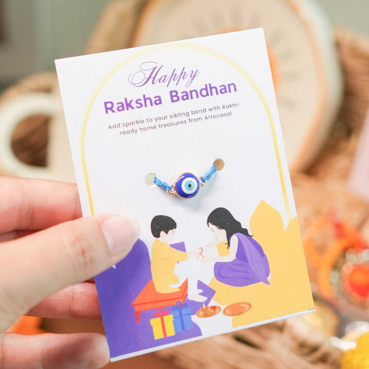 Personalized Handmade Evil Eye Design Guardians Blessing Rakhi Hamper Includes 1 Basket 1 Cup And Saucer 1 Rakhi 1 Roli Chawal And 1 Message Card - Hand showing rakhi card