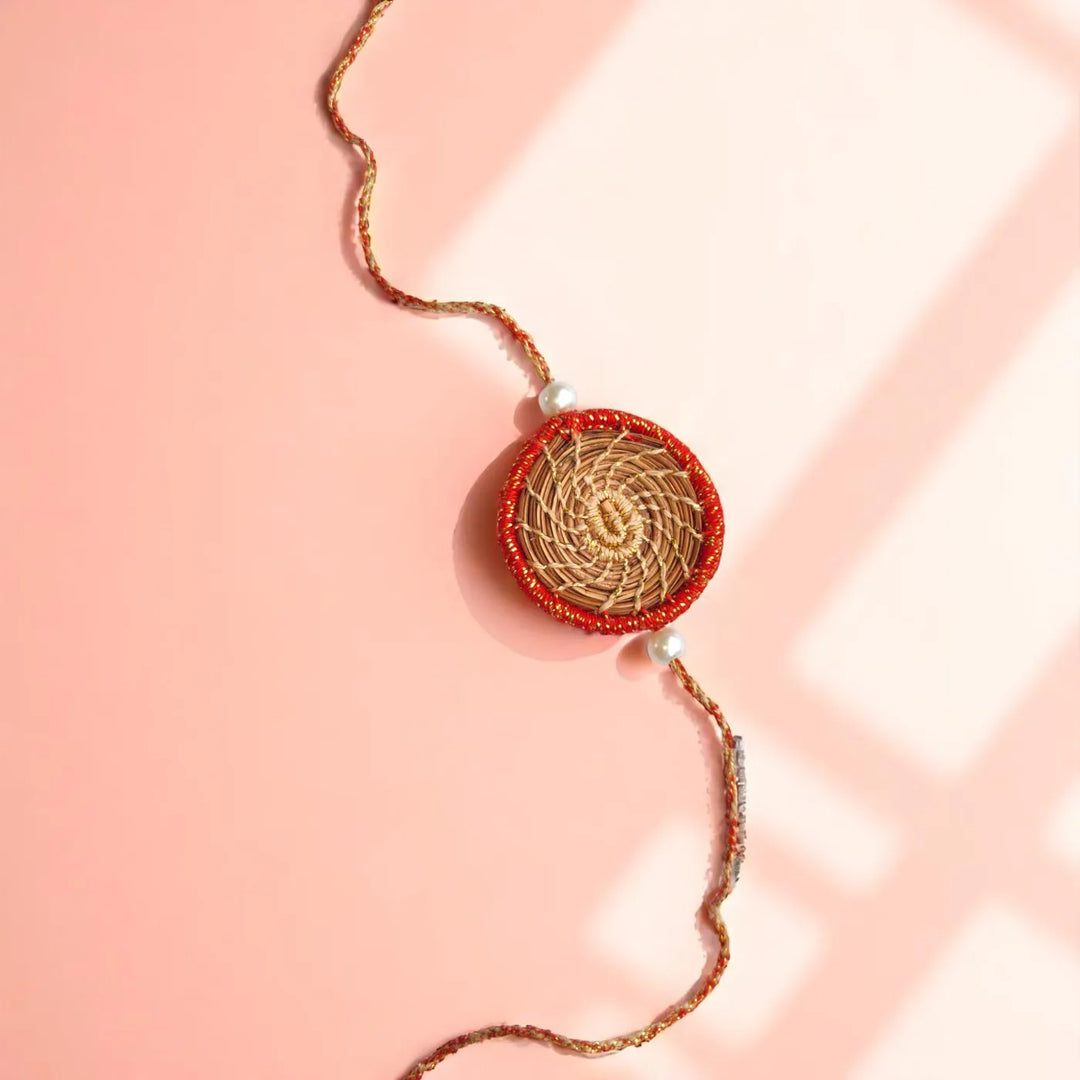 Eco Friendly Handmade Gold And Red Pine Needle Rakhi With Pouch And Roli Chawal - Flat Lay on Pink Background