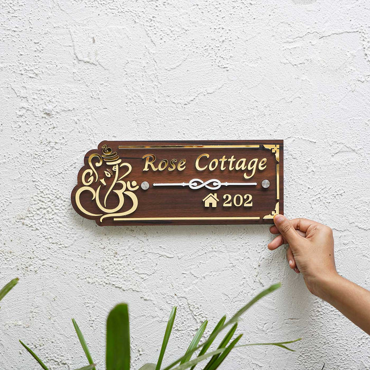 Personalized Weatherproof Brown And Gold Ganesh Themed Acrylic Name Plate - Hand Holding Nameplate Against Wall
